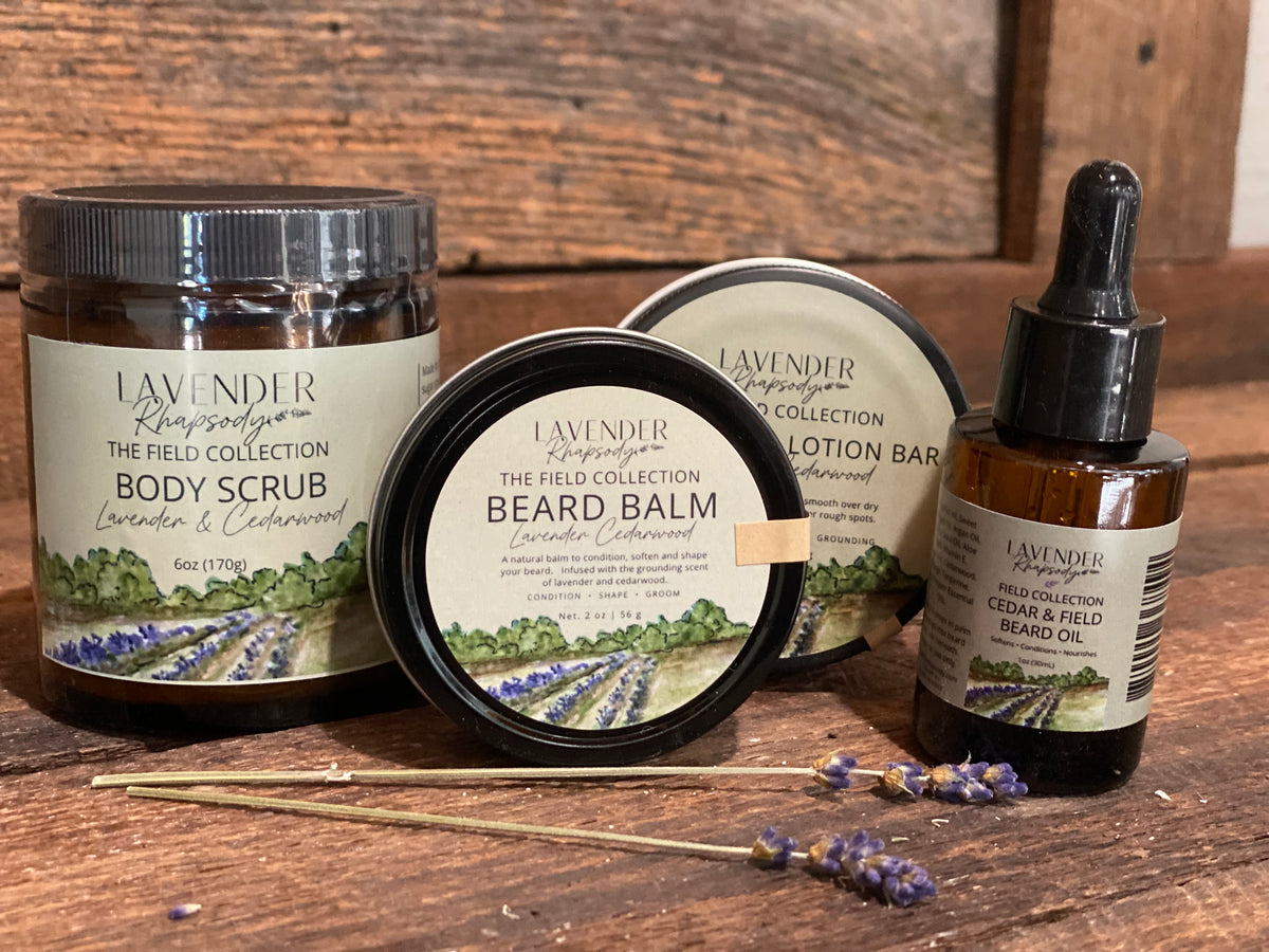 Four lavender-themed skincare and grooming products from Lavender Fields The Field Collection arranged on a rustic wooden surface: a body scrub jar, a beard balm tin, a lotion bar tin, and a beard oil dropper bottle, with lavender sprigs in front.