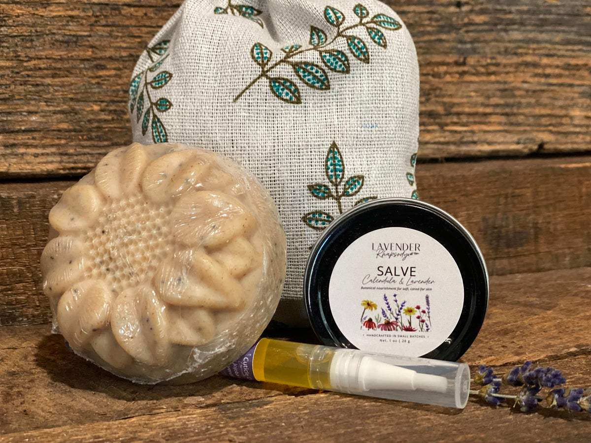 A bath and body gift set on a rustic wooden background, featuring a sunflower-shaped bar of soap in plastic wrap, a small spray bottle, a tin of Lavender Farm Salve, and a beige linen drawstring bag with green and blue leaf embroidery.