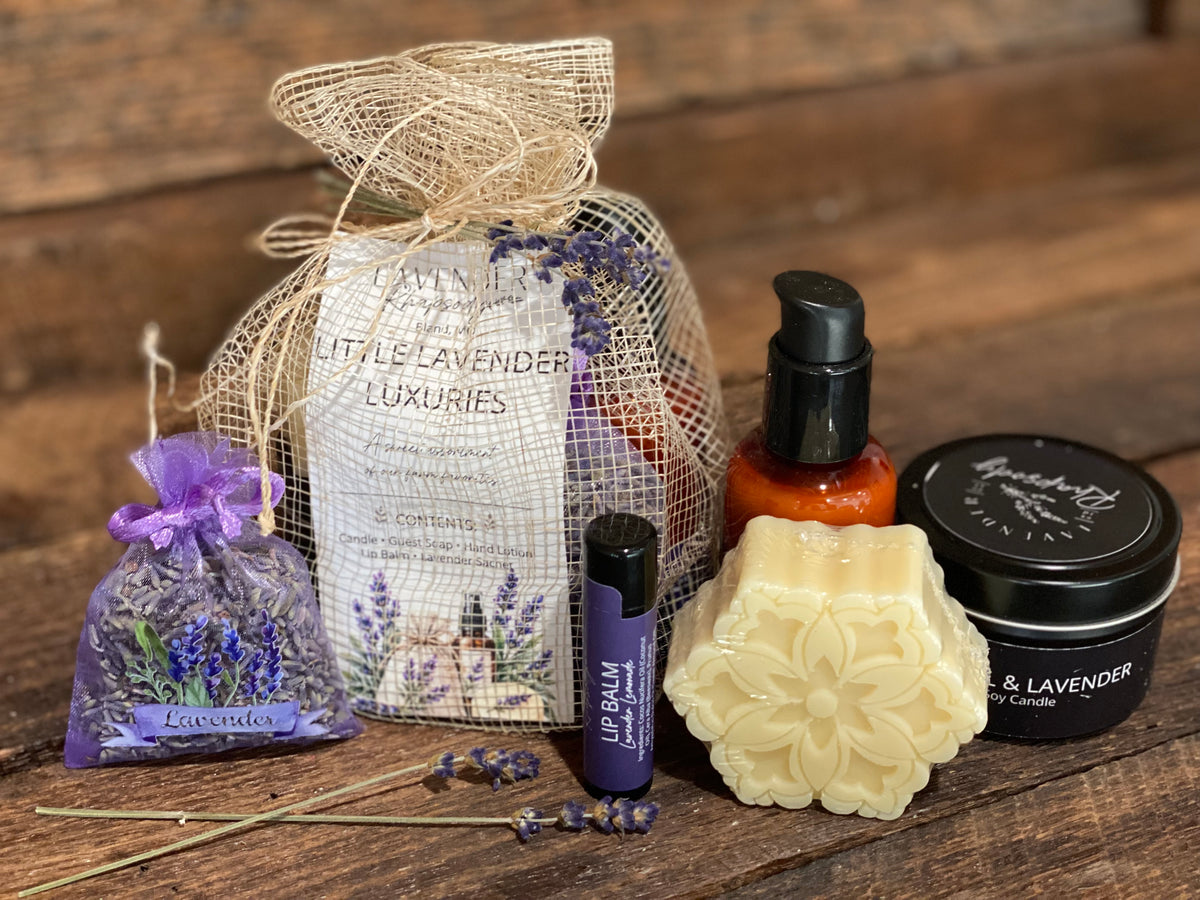 A lavender-themed gift set arranged on a rustic wooden surface, including a lavender sachet in a mesh bag labeled 'Little Lavender Luxuries,' a purple lip balm tube, a flower-shaped cream soap bar, a black candle tin, and an amber bottle.
